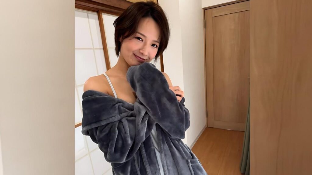 Ai Mukai with short dark hair in a gray plush robe draped off her shoulders, white strap top visible, wooden door.
