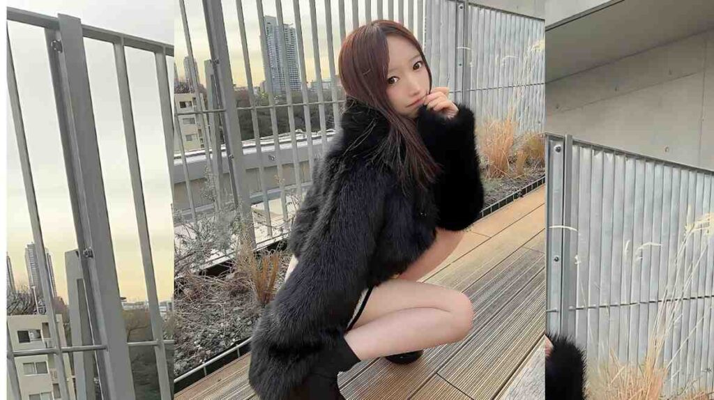 Airi Shimizu in a dark fur coat crouches on a wooden rooftop deck, hand near her mouth, with city skyline in the distance.