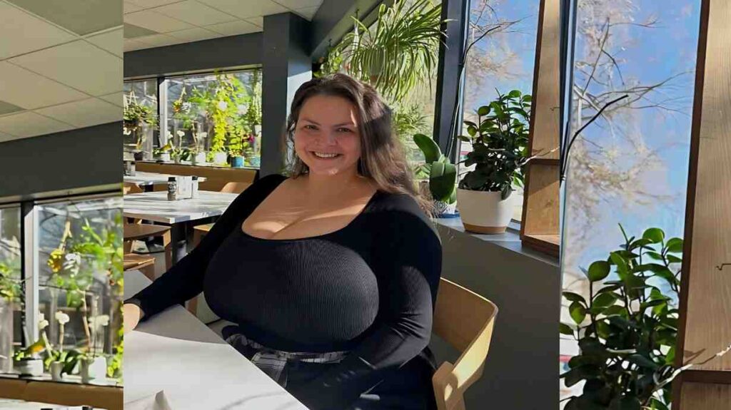 Alaura Grey with long dark hair sits at a sunlit cafe table by a window, beside potted plants, wearing a black ribbed top.