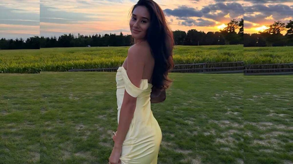 Alex Adams in a pale yellow off-shoulder dress, turned toward the camera with long dark hair flowing.