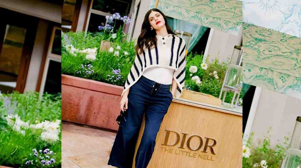 Alexandra Daddario in white top and striped poncho, navy wide-leg pants, posing beside a Dior display outdoors among blossoms.