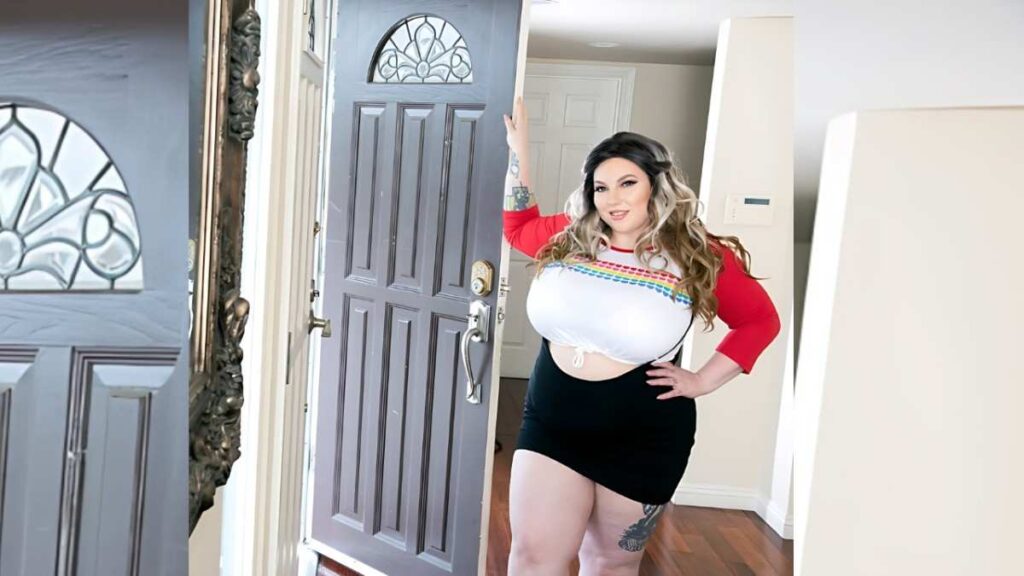 Amy Villainous with wavy blonde hair stands in doorway, wearing a red top, cropped tee in rainbow trim, black mini skirt.