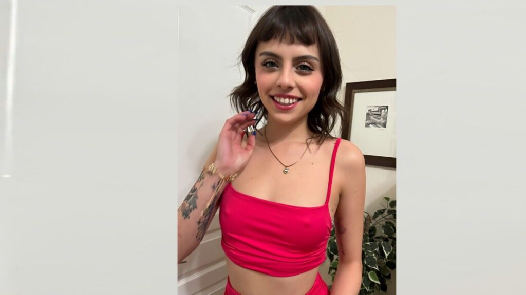 Angel Windell with short dark hair and bangs wears a bright pink cropped top, showing a tattooed left arm and a necklace.