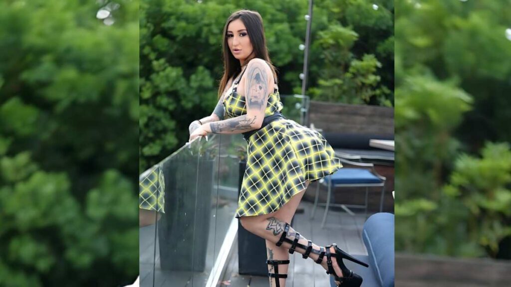 Anna Chambers in a yellow plaid dress leans on a glass railing, tattoos on arms and leg visible, wearing black strappy heels, with greenery and outdoor seating behind.