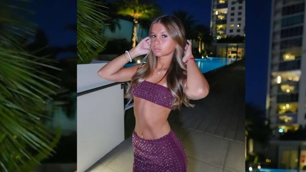 Ashley Rose in a sparkly purple two-piece stands by a rooftop pool at night, touching her wavy hair with city lights behind.
