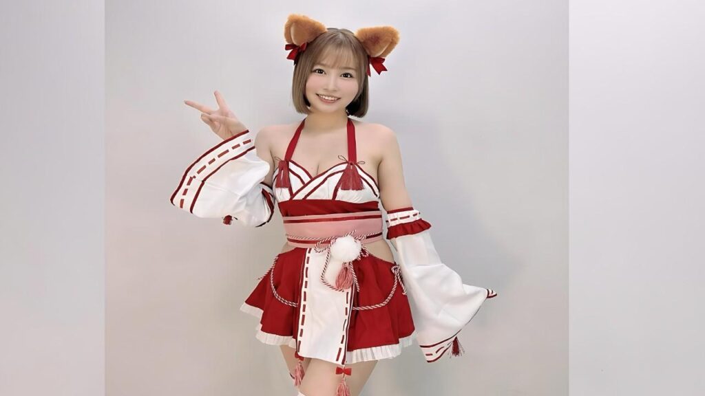 Asuna Kawai in red-and-white outfit with cat ears and tassels, posing with a peace sign against a plain wall.