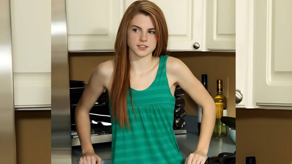 Aubrey Belle with long red hair in a green striped sleeveless top leans on a kitchen counter, white cabinets behind her.