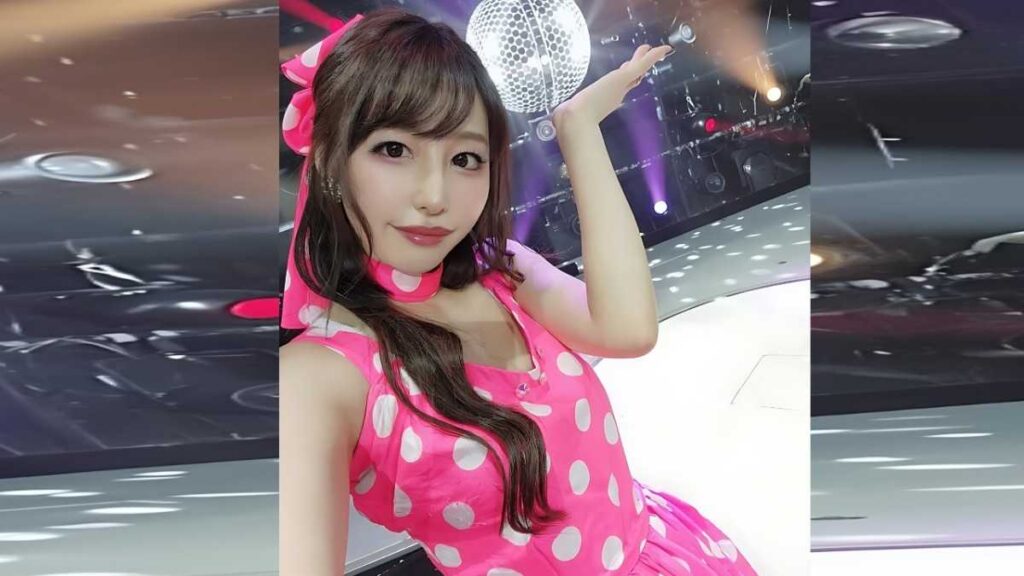 Ayami Shunka in a pink polka-dot dress and matching bow poses with a raised hand, against a stage backdrop with disco ball.