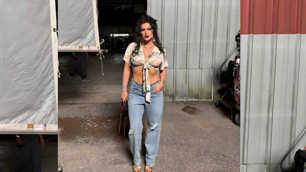 Bella Thorne stands in an industrial yard, wearing a beige tied crop top and ripped jeans, holding a small bag.