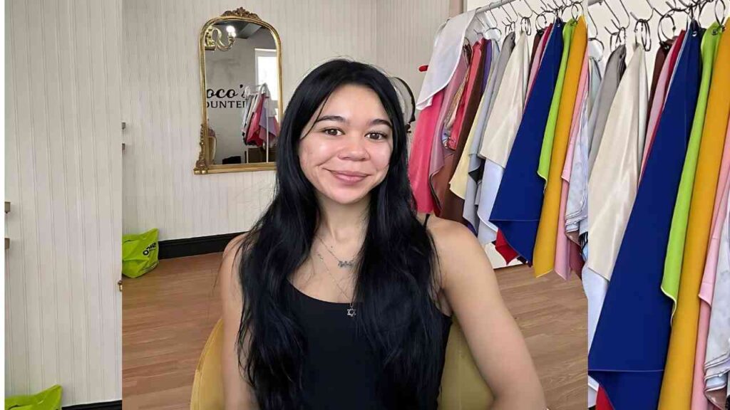Brianna Arson with long black hair and a black tank top sits in a boutique, clothing on racks and a gold-framed mirror above.