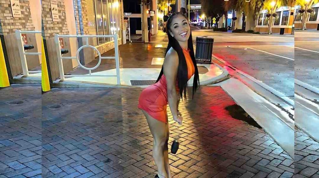 Camila Cortez in orange top and pink shorts leans forward on a sidewalk, wearing cat-ear headband and holding a black purse.