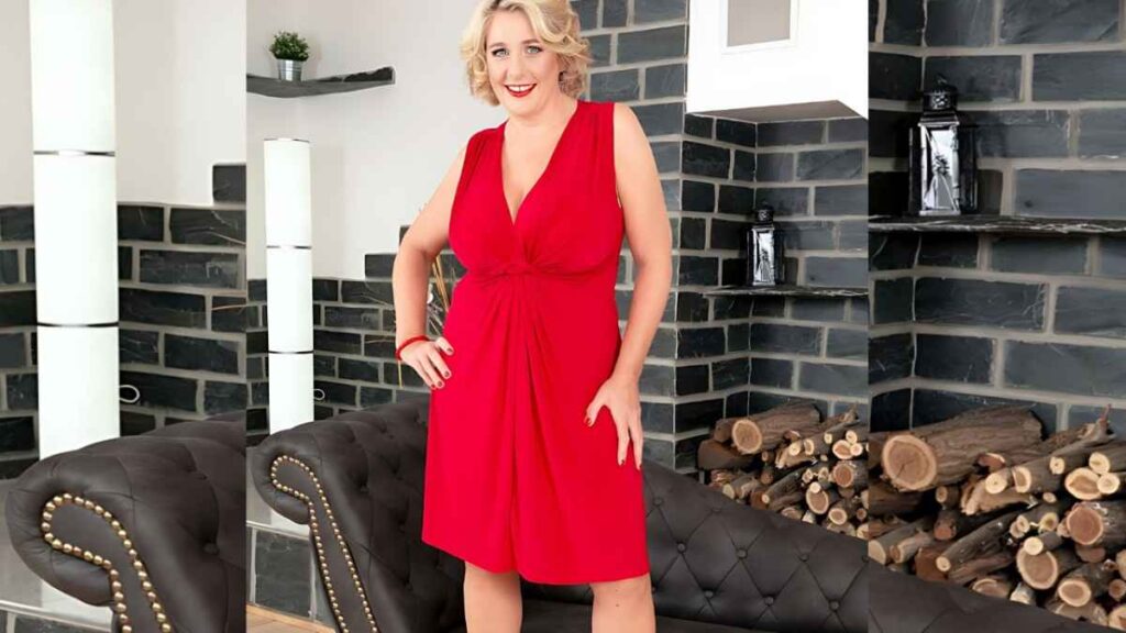 Camilla Creampie in a red knee-length dress stands with hands on hips in a chic room with black stone walls and sofa.