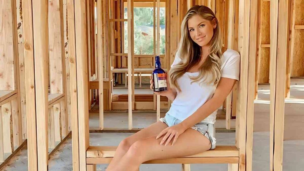 Carly Lauren in a white tee and denim shorts sits on a wooden bench inside a framed construction site, holding blue bottle.