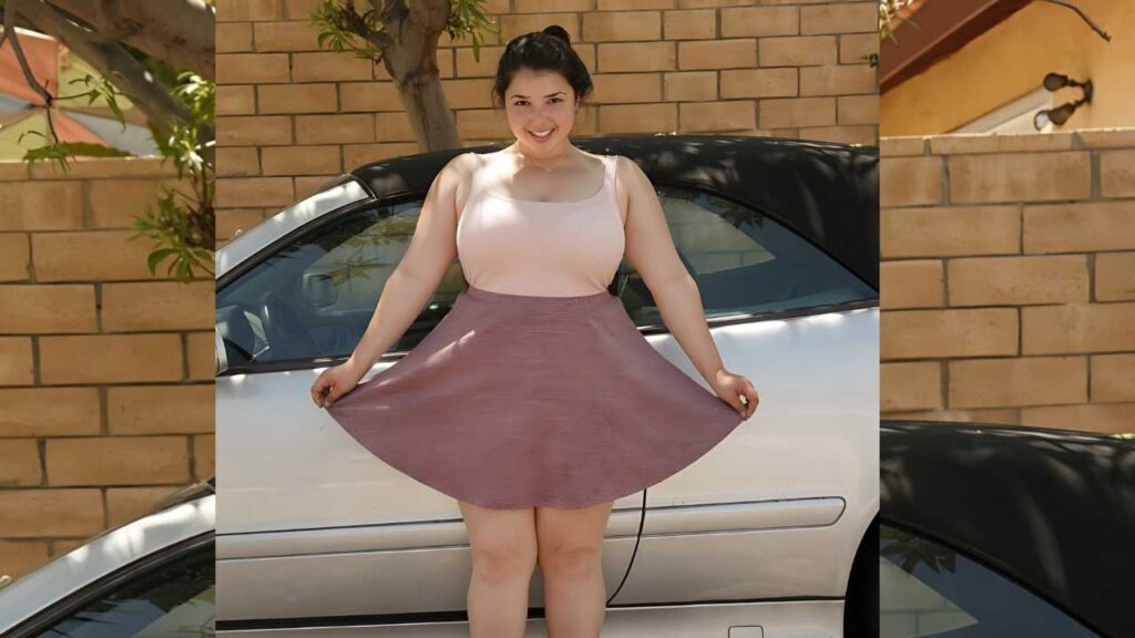 Carolina Munoz in a pale pink tank top and mauve swing skirt holds the skirt out beside a silver car, brick wall behind.
