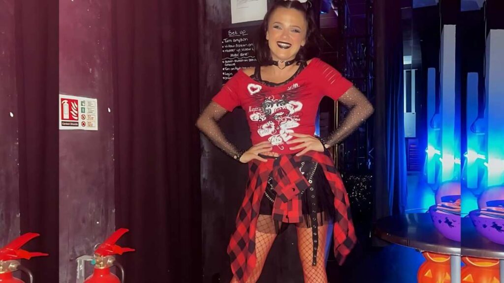 Charley Hart in red graphic tee, fishnet tights and plaid shirt tied at the waist, posing with hands on hips backstage.