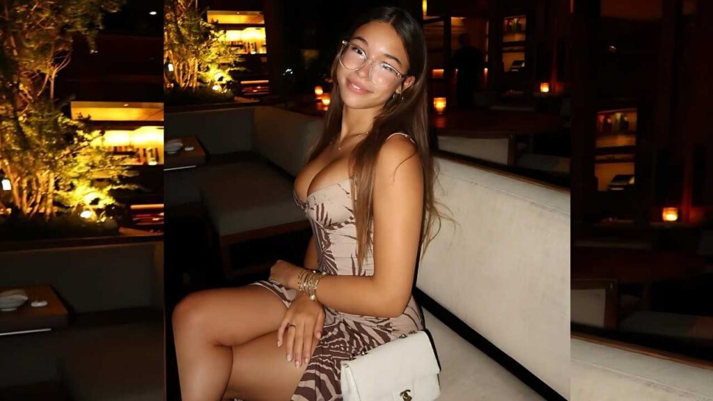 Chloe Parker with long hair and glasses sits on a cream couch in a dim lounge, wearing a patterned dress and a white purse.