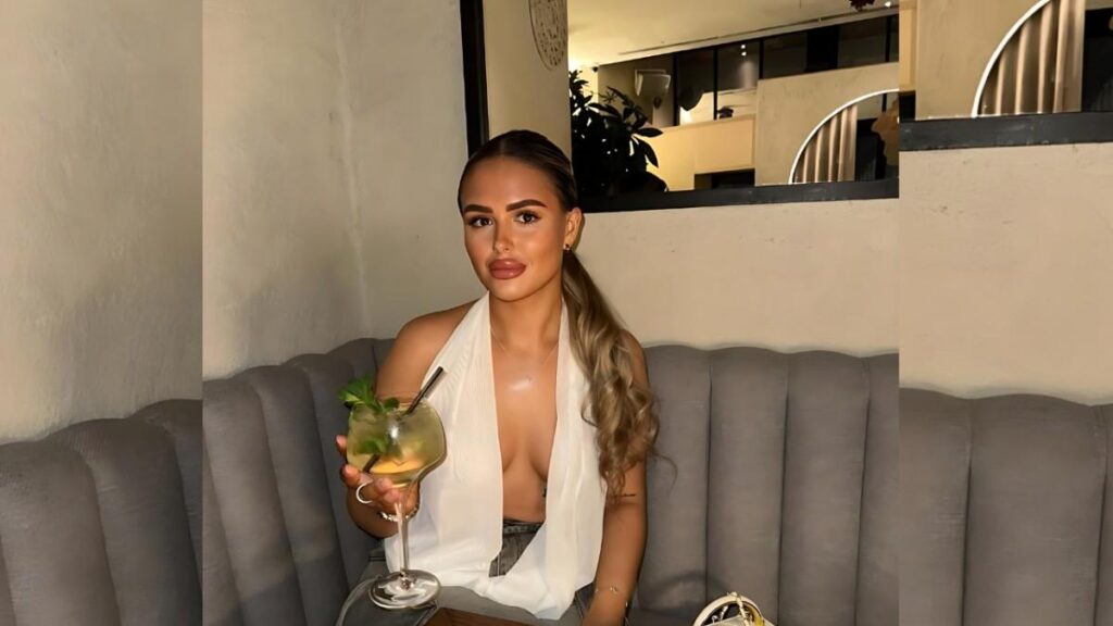 Chloe Scott with a long wavy ponytail sits on a gray tufted banquette, wearing a white halter top and sipping a mint cocktail.