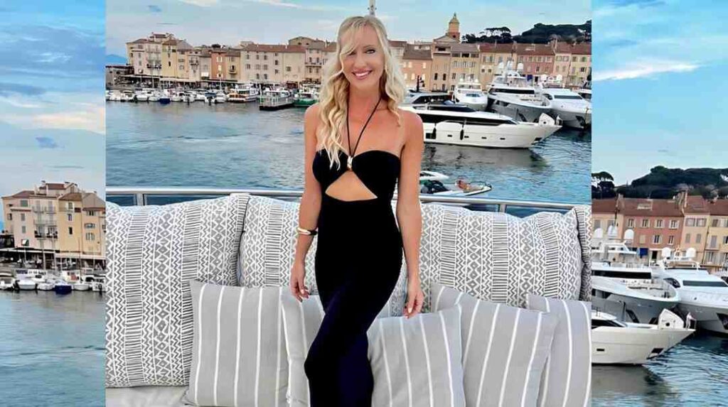 Cindy Shine with long, wavy hair in a black cut-out halter dress poses on a yacht deck, with marina and pastel town behind.