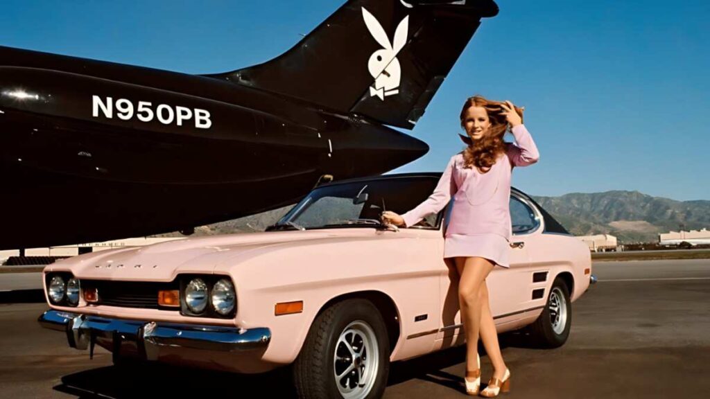 Claudia Jennings in a pink dress poses beside a vintage pastel car on a desert tarmac, with black jet tail featuring white Playboy bunny logo.