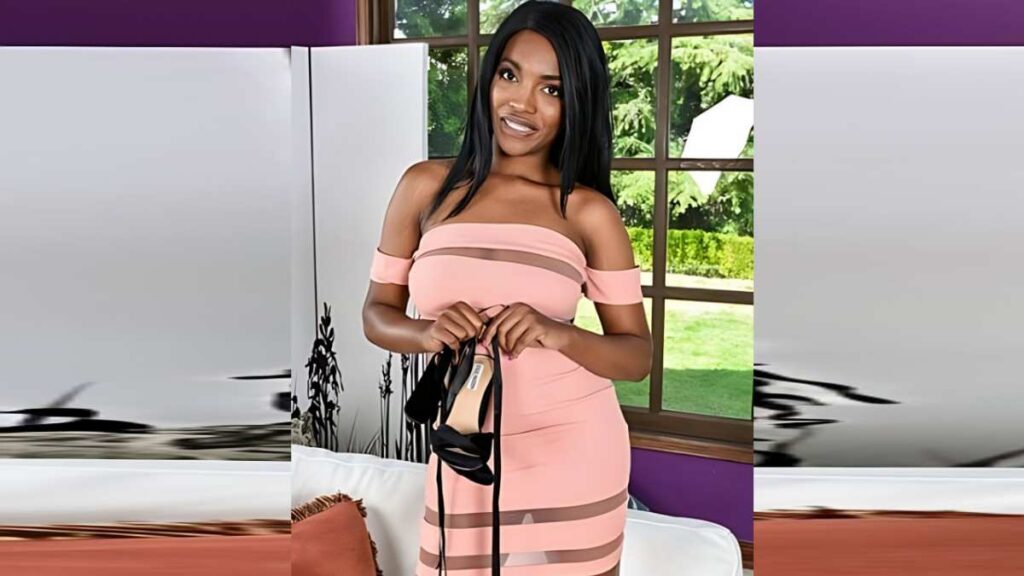 Daya Knight in a pink off-shoulder banded dress holds black high heels, standing beside a purple wall and greenery outside.