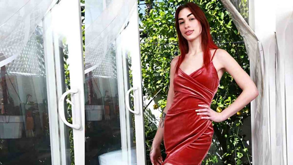 Delilah Day in a red velvet dress poses with hand on hip beside sliding glass doors, lush green foliage behind.