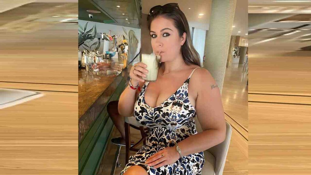 Denise Davies in a blue-and-white floral dress sits at a bar, sipping a pale milkshake through a straw, with taps and greenery behind her in a bright cafe.