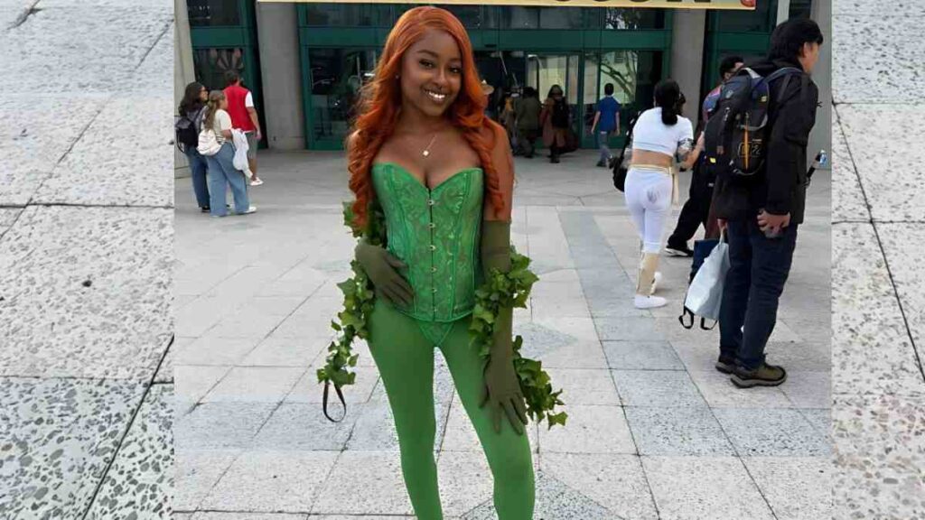 Destiny Mira with long orange hair in a green corset and leggings, ivy garlands around her arms, posing in a busy plaza.