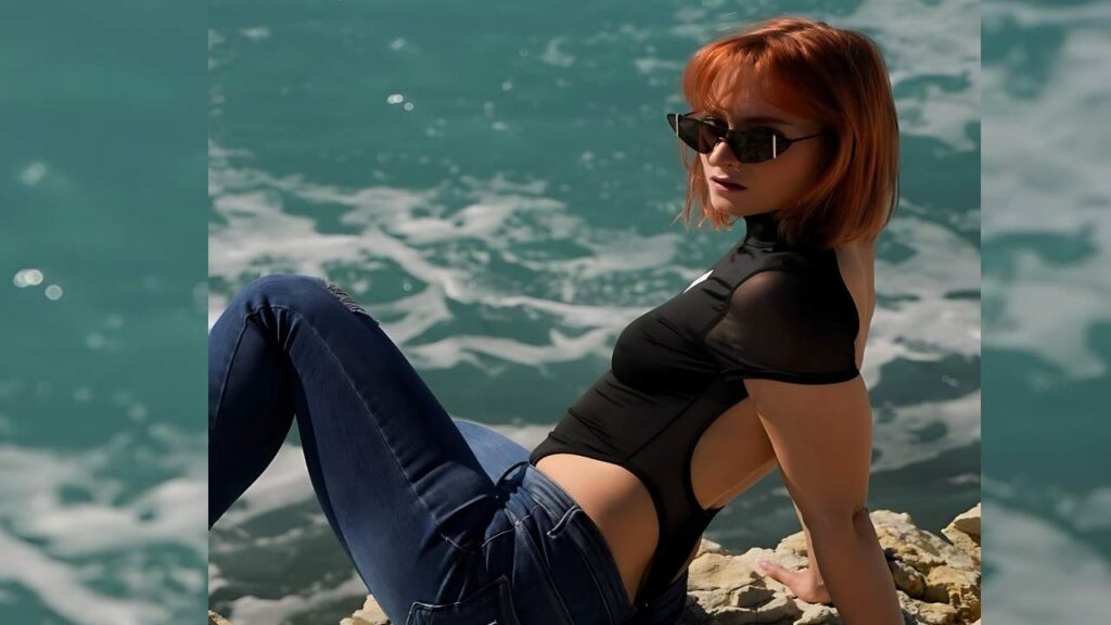 Dolly Dyson with orange bob in black sheer bodysuit and dark jeans sits on rocky ledge beside turquoise water, wearing sunglasses.