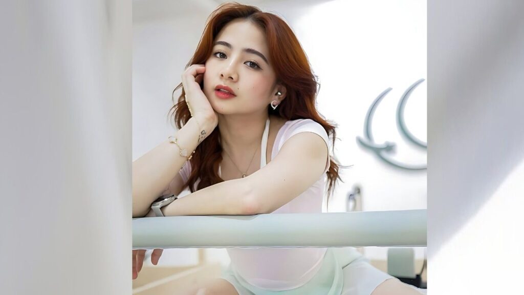 Ella Cruz with auburn hair leans over a pale railing, chin in hand, wearing a light pink top and delicate jewelry.