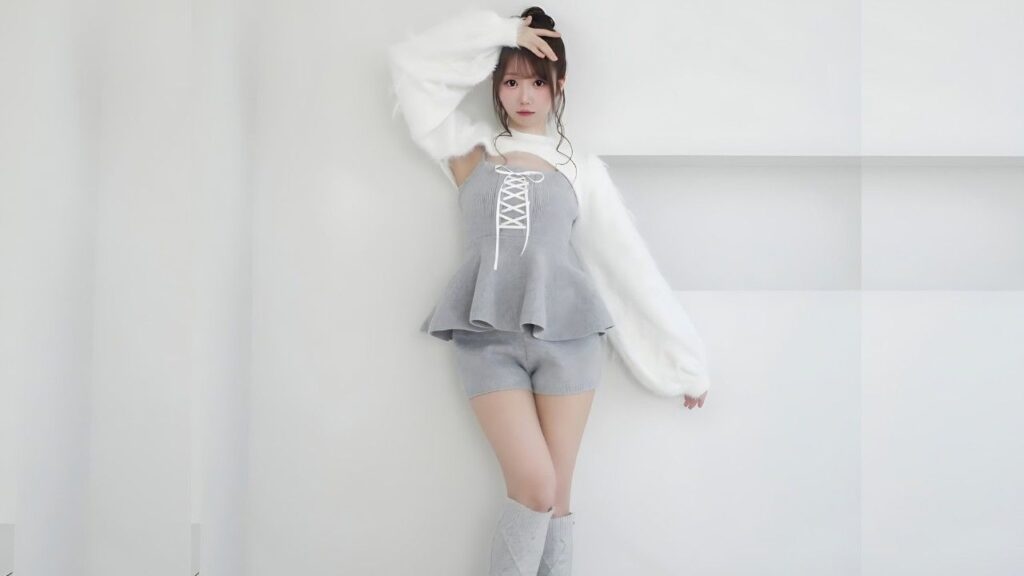 Emiri Ōtani poses against a white studio wall, wearing a grey lace-up top with matching shorts and a fluffy white cardigan, one hand in her hair.