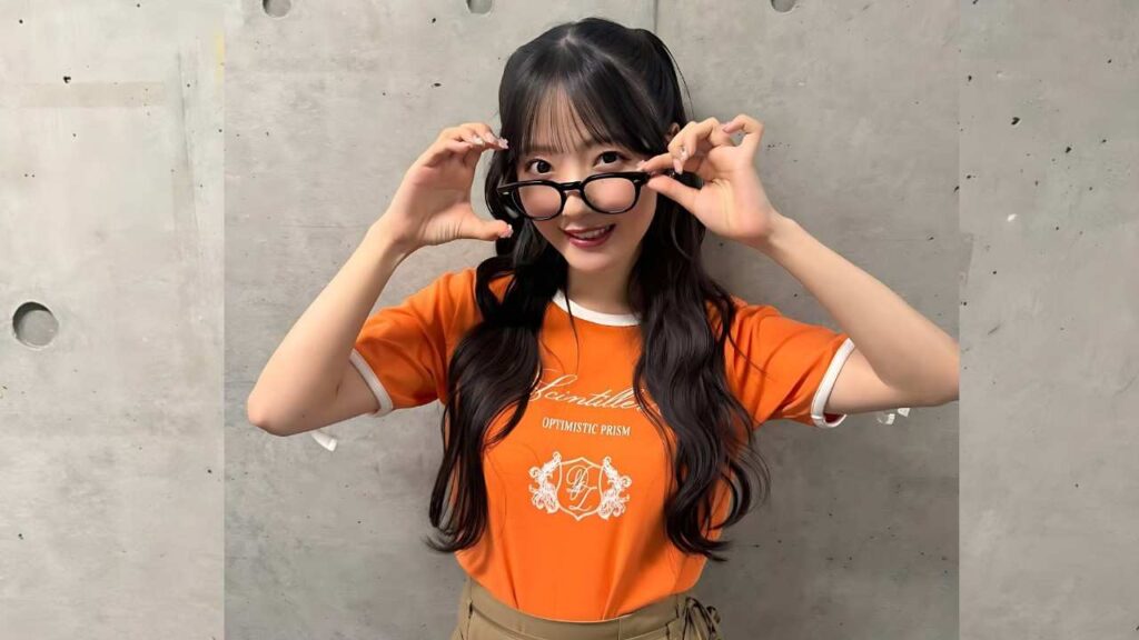 Emiru Kawamoto with long black hair in an orange T-shirt holds glasses and smiles against a gray concrete wall.