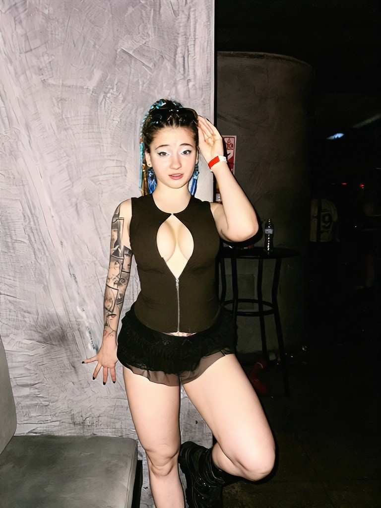Emma Fiore Blue-dyed hair, black zip-front top, lace mini skirt; tattoos on left arm; she poses with hand on head before a club wall.