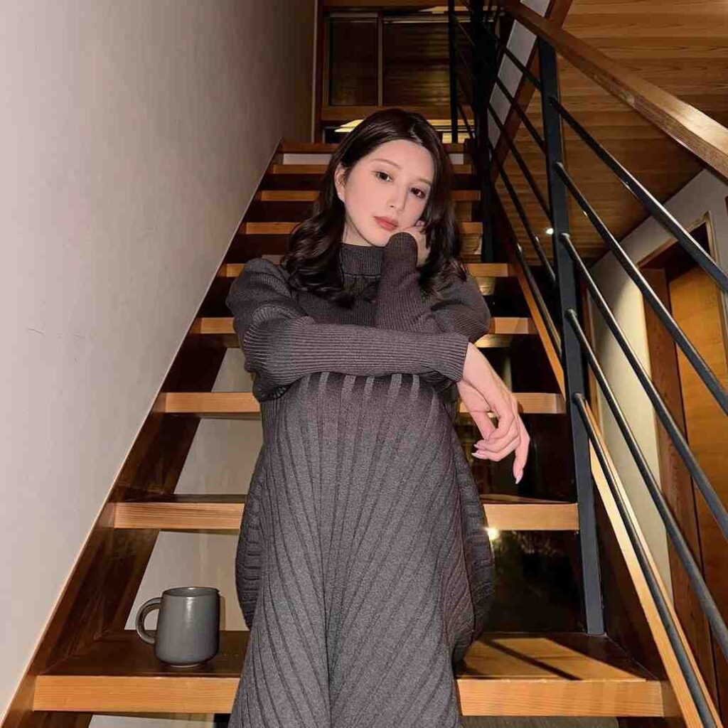 Hikaru Nagi on a wooden staircase wears a gray knit dress, resting her chin on her hand, with a gray mug nearby.