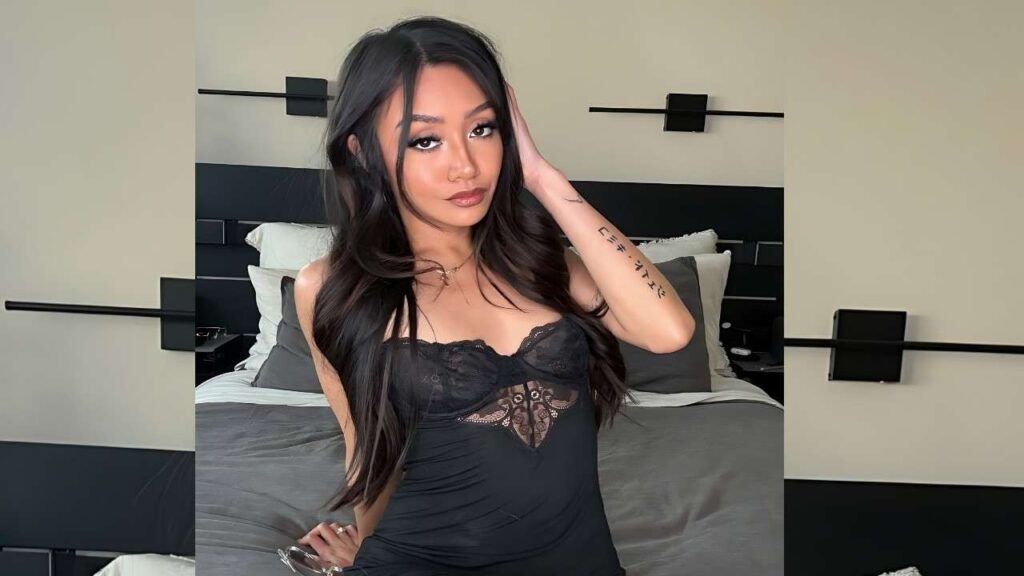 Jade Kimiko in a black lace camisole sits on a gray bed in a modern room, hand in hair, tattooed forearm visible.