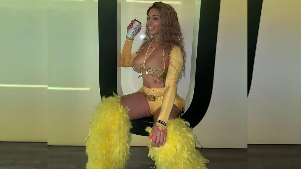 Jasmine Lotus in a yellow bikini-style outfit with feathered yellow leg warmers sits on a chair, holding a Nutril can.