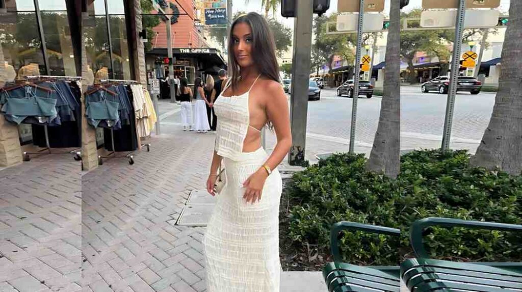 Jenna Rose in a white two-piece set poses on a sidewalk outside shops, with clothing racks, palm trees, and a street in view.