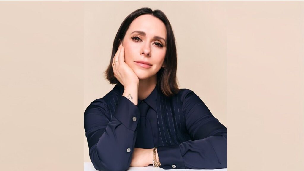 Jennifer Love Hewitt with dark shoulder-length hair in a navy pinstripe blouse, resting her chin on her hand.