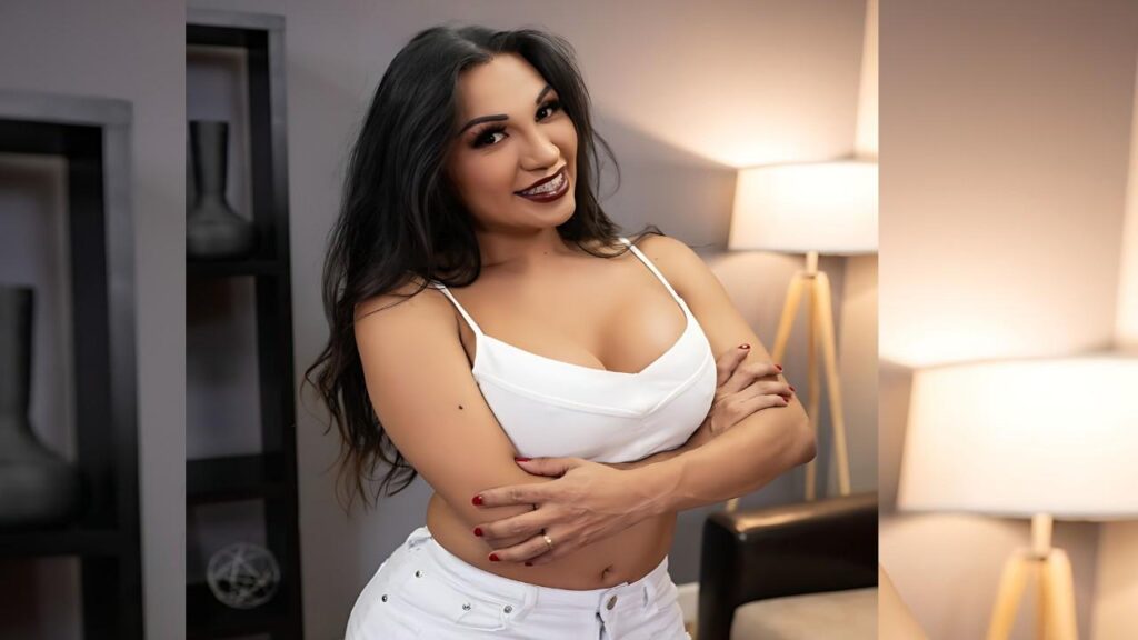 Jessy Dubai with long dark hair in a white top and jeans crosses her arms in a cozy, modern living room with warm lamps.