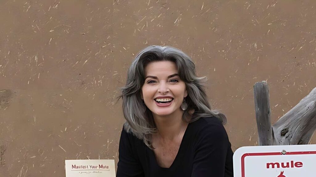 Joan Severance with shoulder-length gray hair in a black top, outdoors against a tan textured wall with a wooden fence.