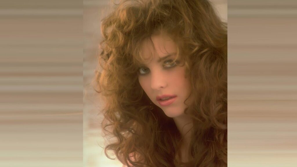 Julia Parton with voluminous curly red hair, smoky eyes, pink lips, gazing at the camera.