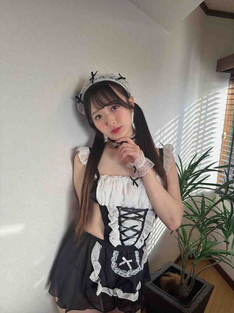 Kaho Hanamori in black-and-white maid cosplay with lace headband, pigtails, and gloves, posing by a wall beside a potted plant.
