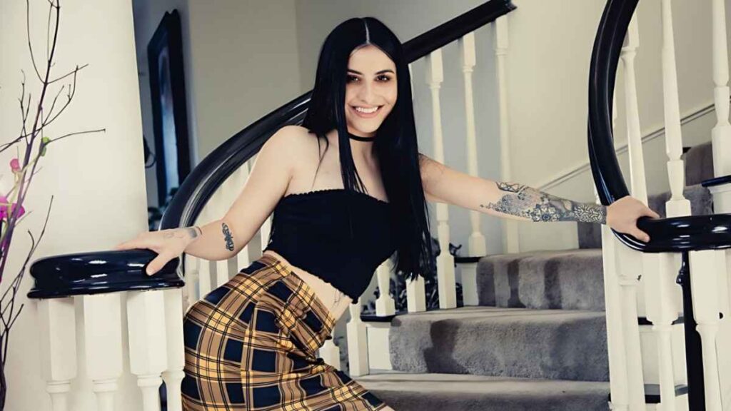 Kaitlyn Katsaros with long black hair leans on a white staircase railing, wearing a black tube top and yellow plaid skirt.