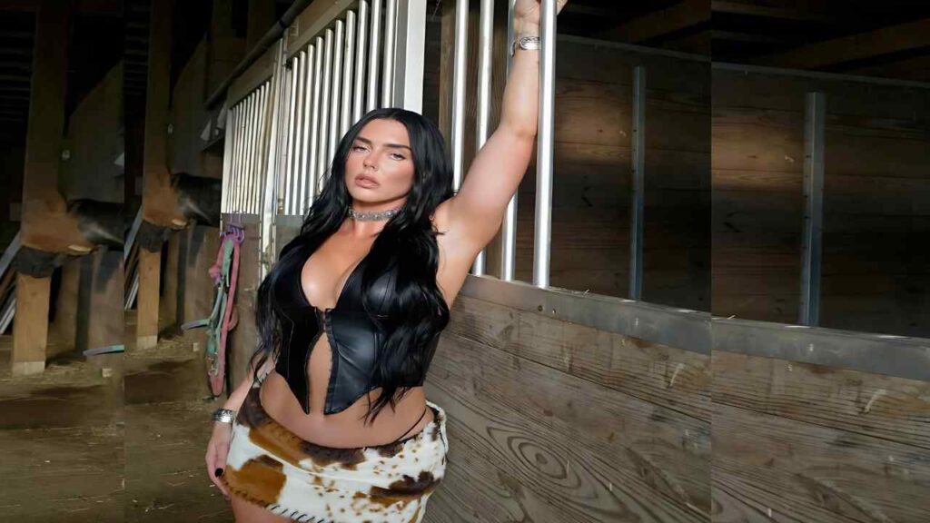 Kaitlynn Anderson with long black hair in a black leather corset and cowhide skirt stands in a barn, arm raised on metal bars.