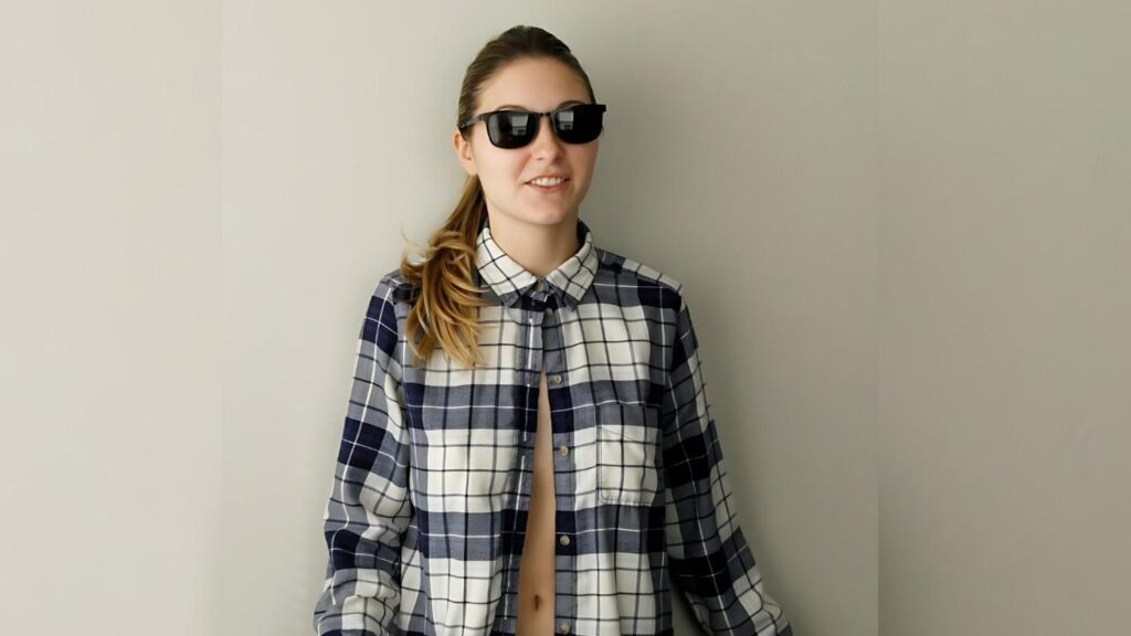 Katie Darling in dark sunglasses wearing a blue-white plaid shirt, hair in a side ponytail, shirt unbuttoned.