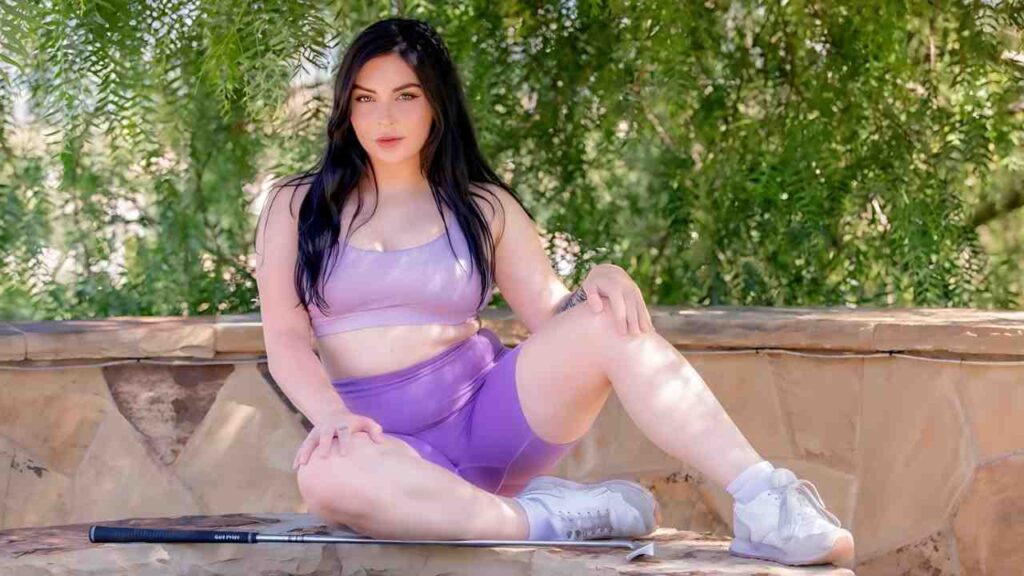 Keira Croft with long dark hair in lavender athletic wear sits on a stone ledge outdoors, a golf club beside her, lush green foliage behind.