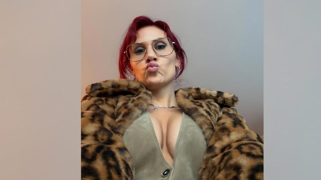 Kelli Fire in aviator glasses and leopard-print fur coat, plunging green top, pursed lips in a selfie.