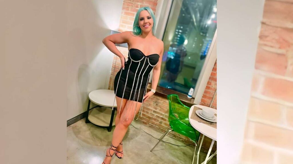 Lady Dee in a black strapless mini dress with silver fringe, posing with hand on hip in a brick-walled room.