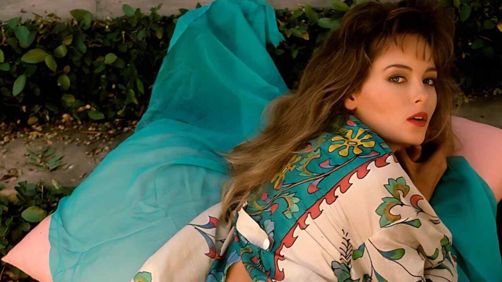 Lisa Matthews lies on a pink cushion outdoors, wrapped in teal and a colorful floral shawl, gazing at the camera.