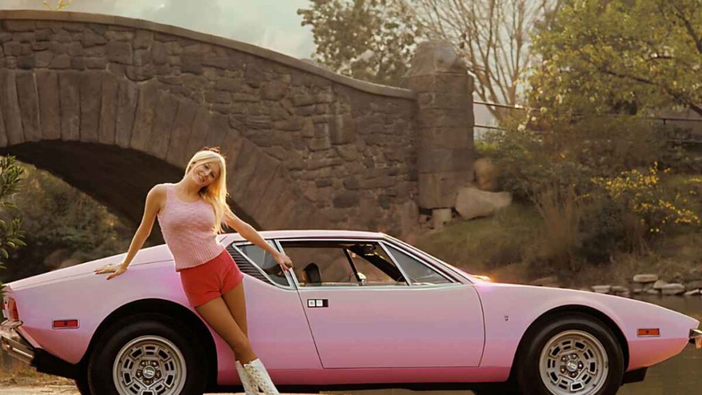 Liv Lindeland in a pink sleeveless top and red shorts leans on a light pink vintage sports car beside a stone arch bridge.