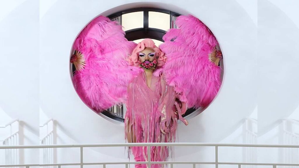 Lola Rose with pink feather wings and shimmering pink gown poses before a circular window, pink wig and bold makeup.